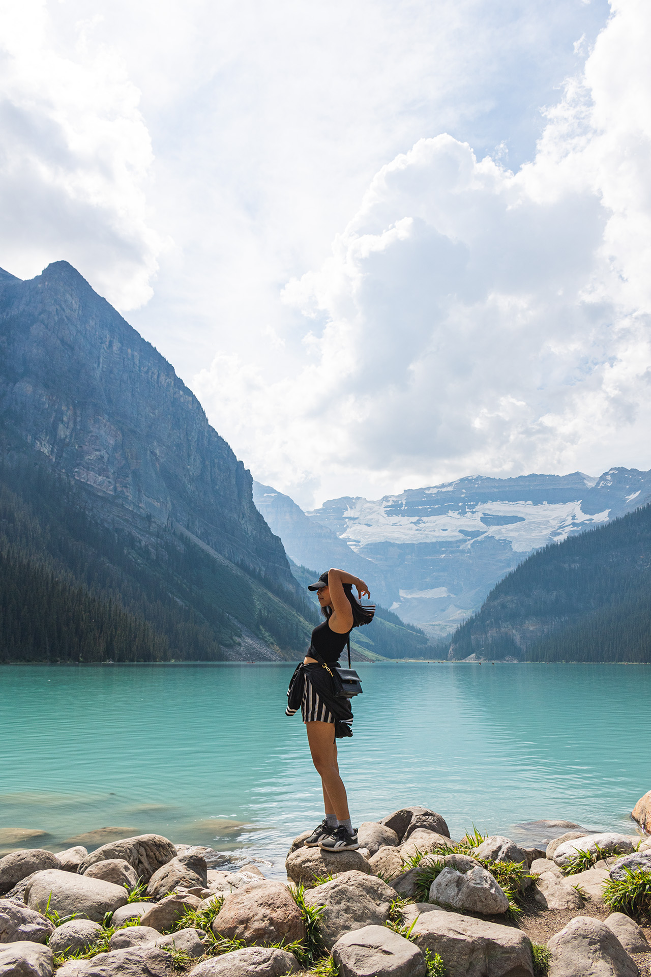 Girl flipping her hair as she stands on a stone near a teal blue lake behind her, surrounded by rocky mountains.
